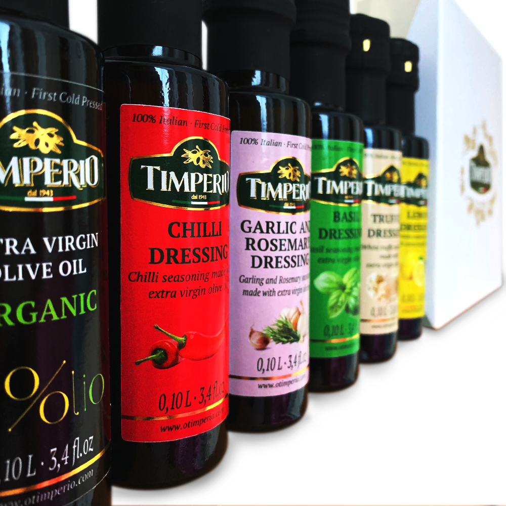 A row of Timperio Italian virgin olive oil and dressing bottles in different flavors, including Chilli Dressing, Garlic and Rosemary Dressing, Basil Dressing, Truffle Dressing, and Extra Virgin Olive Oil, shown alongside a white branded gift box.