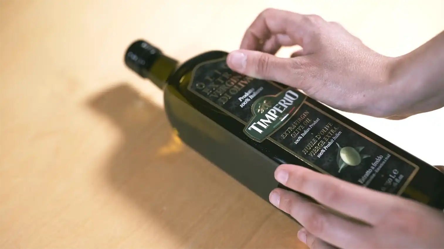 A person's hands holding a bottle of Timperio Oliva Extra Virgin Olive Oil on a light wooden surface.